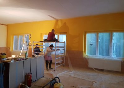 A group of volunteers is renovating the colorful Kalapa Tibetan Buddhist center. The space has bold yellow walls, with a man and two women actively painting using various tools and workbenches. The room contains construction materials, a ladder, and floor coverings for protection.