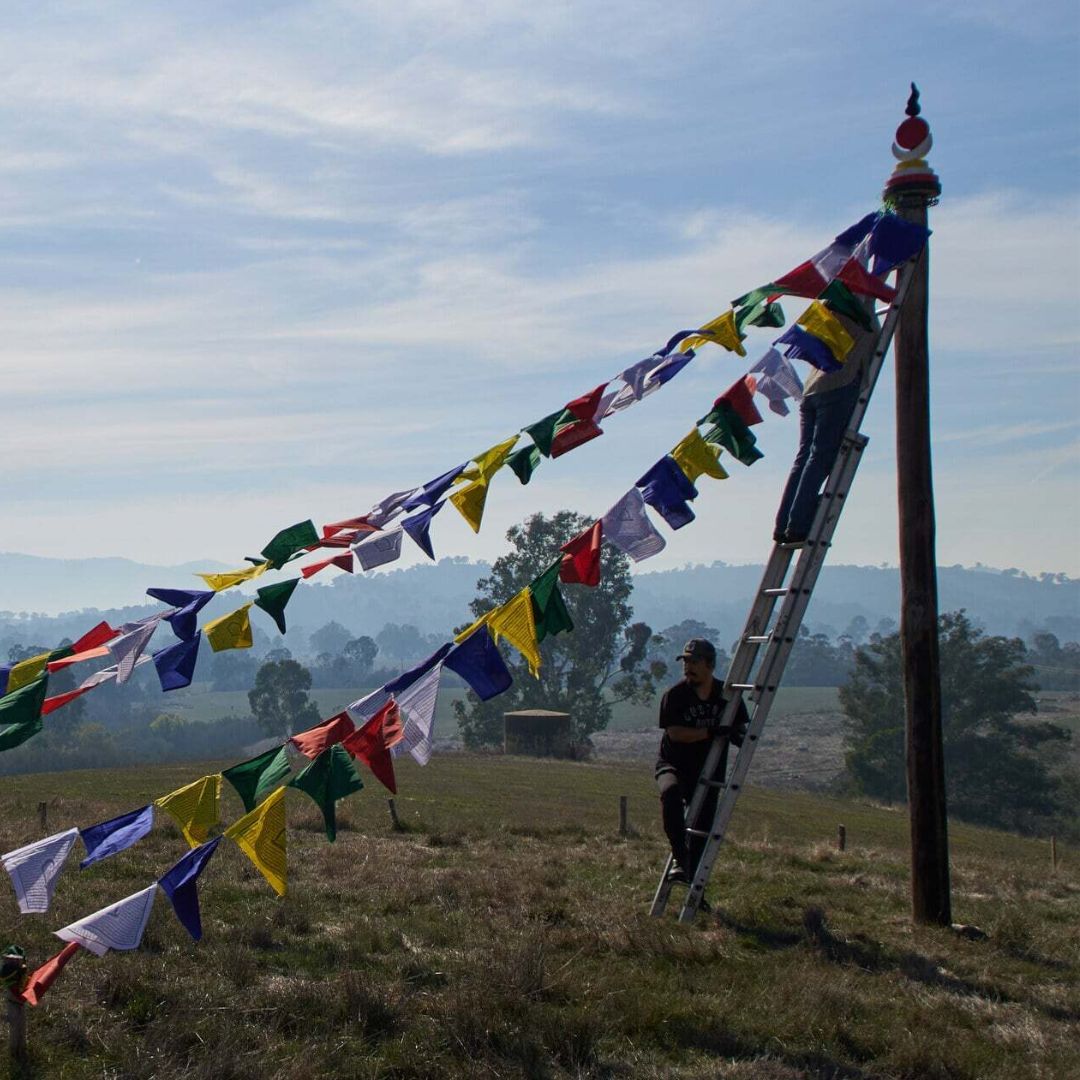 volunteers raising tibetan prayer flags on Saka Dawa at Land of Shambhala. One is on a ladder and the other is helping the person connect the flags to the pole that has a sun moon rahu and kalgni disk on it with a nada rising to the sky. 